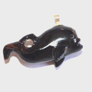 May include: A dark brown dolphin pendant with a gold-colored bail. The pendant is carved with a smooth, glossy finish, featuring a detailed dolphin shape with fins and a smiling mouth. The dolphin is approximately 2 inches long.