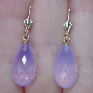 May include: A pair of gold-toned drop earrings featuring faceted, teardrop-shaped lavender gemstones. The earrings have a lever-back closure and are displayed against a blurred background.