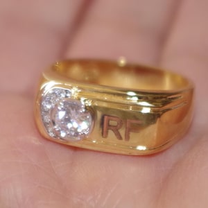 Könnte beinhalten: Ein goldfarbener Ring mit einem großen, klaren, runden Edelstein, der in einer Ansammlung kleinerer Steine gefasst ist. Der Ring hat ein rechteckiges Band mit den Initialen "RF" eingraviert.