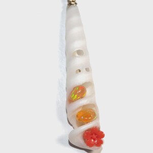 May include: A long, white, spiral-shaped shell pendant featuring a gold-toned metal bail at the top. The shell is adorned with two small, round, iridescent orange opals and a single, carved, bright red flower at the bottom opening.