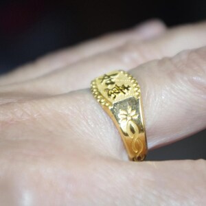 May include: A gold ring with an engraved design and Chinese characters. The ring is on a finger, showcasing its intricate details. The ring has a floral design on the side.
