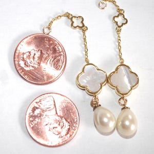 May include: Gold-toned drop earrings featuring a four-leaf clover design with a mother-of-pearl inlay and a teardrop pearl. The earrings are shown next to two pennies for size comparison. The earrings are on a white background.