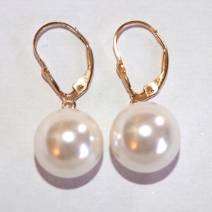 Puede incluir: Un par de pendientes dorados con cierre de palanca, cada uno con una perla grande y redonda de color crema. Los pendientes se muestran sobre un fondo blanco, resaltando el contraste entre el metal dorado y las perlas.