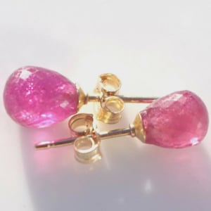 May include: A pair of gold stud earrings with faceted, teardrop-shaped pink gemstones. The earrings have a post and butterfly backing. The gemstones are a vibrant, saturated pink color.