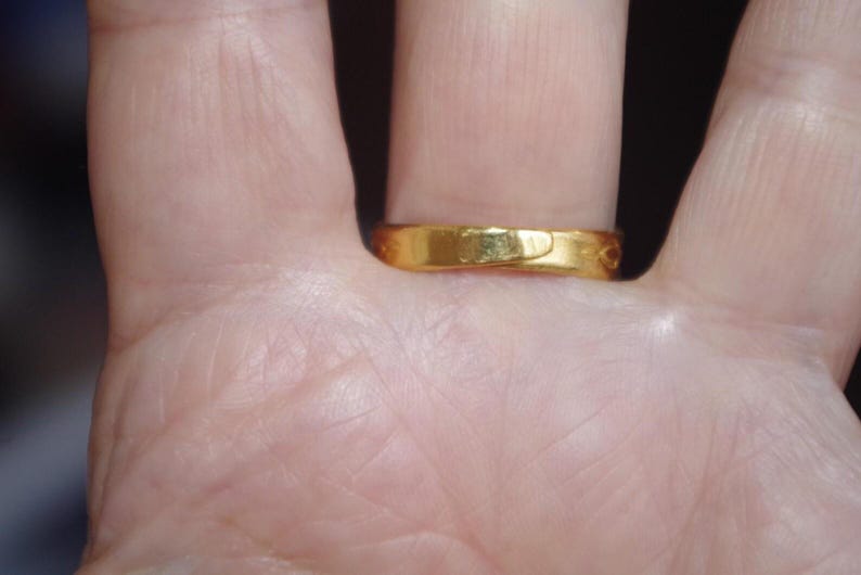 May include: A gold ring with a smooth, polished surface. The ring is worn on a finger, and the close-up shot highlights its simple, elegant design. The ring's warm, golden hue suggests it is made of gold.