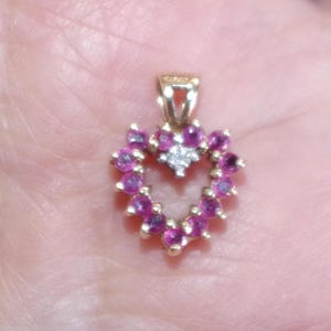 May include: A gold pendant with a heart-shaped design. The pendant is adorned with small, round, pink gemstones encircling a central cluster of white gemstones. The bail is also gold.