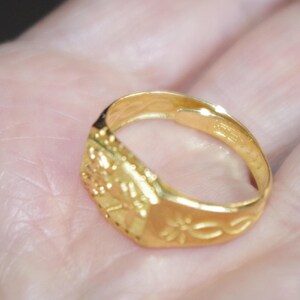 May include: A gold-colored ring with a rectangular face featuring embossed floral and geometric designs. The ring's band has a floral pattern. The ring is held in a hand, showcasing its intricate details and polished finish.