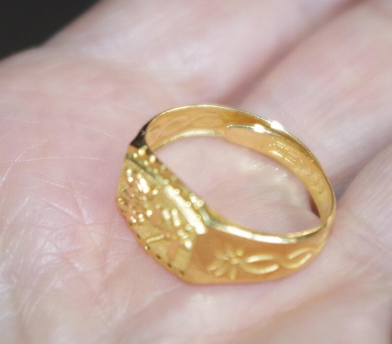 May include: A gold-colored ring with a rectangular face featuring embossed floral and geometric designs. The ring's band has a floral pattern. The ring is held in a hand, showcasing its intricate details and polished finish.