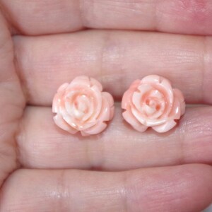 Puede incluir: Un par de pendientes en forma de rosa rosa claro. Los pendientes est&aacute;n tallados con p&eacute;talos detallados y se sostienen en la mano de una persona. Los pendientes son de un delicado tono rosa.