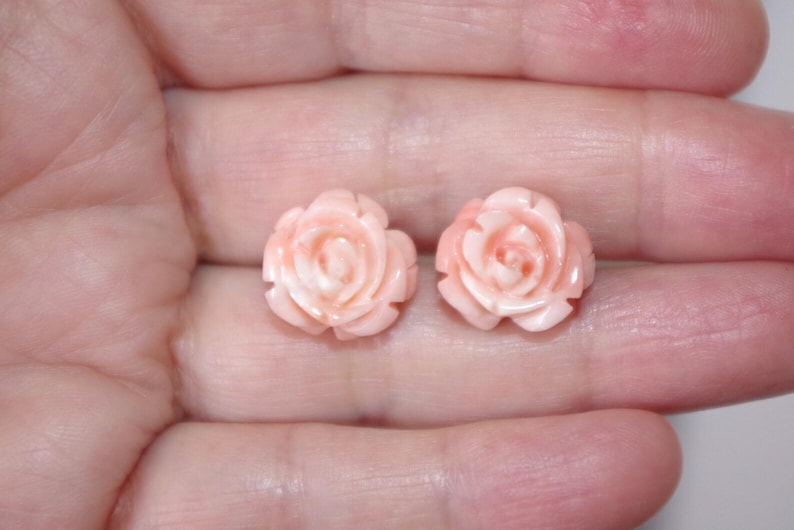 Puede incluir: Un par de pendientes en forma de rosa rosa claro. Los pendientes est&aacute;n tallados con p&eacute;talos detallados y se sostienen en la mano de una persona. Los pendientes son de un delicado tono rosa.