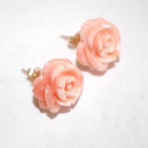 Puede incluir: Un par de pendientes con forma de rosa. Los pendientes est&aacute;n tallados en un material de color coral claro, con intrincados detalles que imitan los p&eacute;talos de una rosa. Los postes son de color dorado. Sobre un fondo blanco.