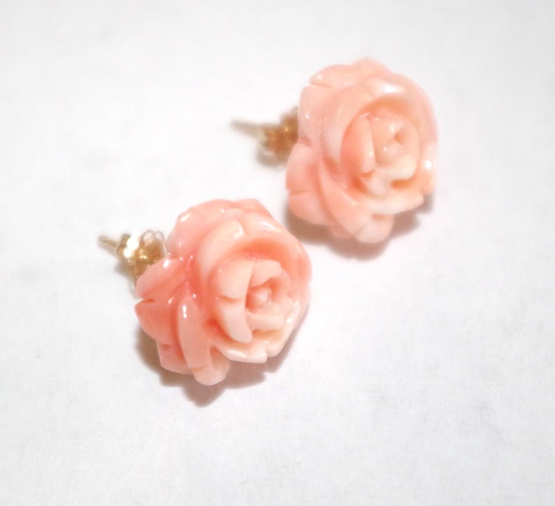 Puede incluir: Un par de pendientes con forma de rosa. Los pendientes est&aacute;n tallados en un material de color coral claro, con intrincados detalles que imitan los p&eacute;talos de una rosa. Los postes son de color dorado. Sobre un fondo blanco.