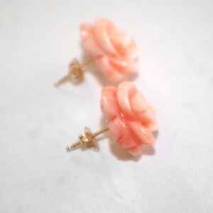 Puede incluir: Un par de pendientes de bot&oacute;n con forma de rosa de color coral. Los pendientes tienen postes dorados y est&aacute;n sobre un fondo blanco. Los p&eacute;talos de rosa est&aacute;n intrincadamente tallados, mostrando la belleza natural del coral.