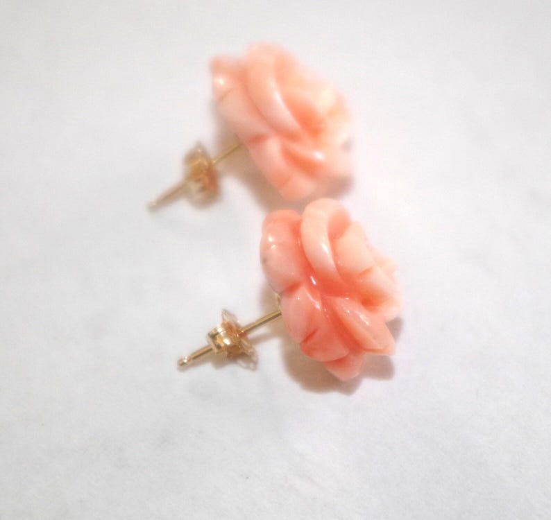 Puede incluir: Un par de pendientes de bot&oacute;n con forma de rosa de color coral. Los pendientes tienen postes dorados y est&aacute;n sobre un fondo blanco. Los p&eacute;talos de rosa est&aacute;n intrincadamente tallados, mostrando la belleza natural del coral.