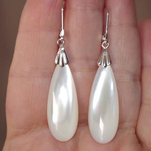 May include: A pair of elegant, pear-shaped earrings. Each earring features a lustrous, off-white, teardrop-shaped pearl suspended from a silver-colored hook. The top of each pearl is capped with a decorative silver element.
