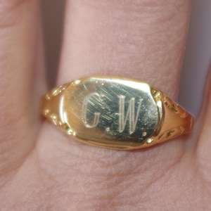 Könnte beinhalten: Ein goldener Siegelring mit den eingravierten Initialen "G.W.". Der Ring hat eine rechteckige Oberfläche mit abgerundeten Kanten und dekorativen Details an den Seiten. Der Ring hat eine warme, glänzende Goldfarbe.