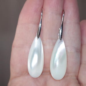 May include: A pair of pear-shaped earrings with a lustrous, white, iridescent surface. The earrings have a silver-colored hook. The teardrop-shaped earrings are displayed against a neutral background.