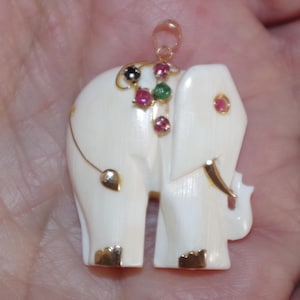 May include: Ivory-colored elephant pendant with gold accents and colorful gemstones. The pendant features a detailed carving of an elephant with gold-plated feet, tusks, and a decorative saddle. The elephant is adorned with red, green, and black gemstones.