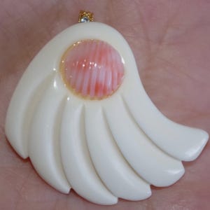 May include: A pendant featuring a smooth, off-white, fan-shaped base with five curved segments. A round, ribbed, light pink coral cabochon is set in the center of the upper portion. A small gold-toned metal bail is attached to the top edge.