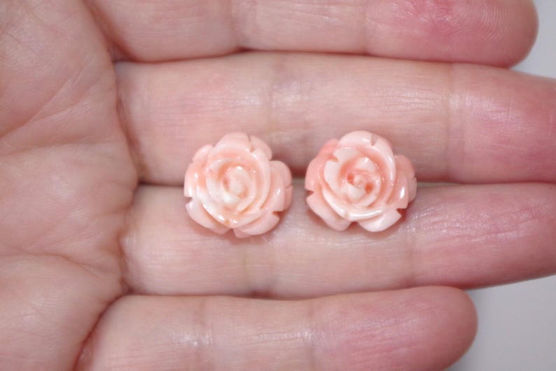 Puede incluir: Un par de pendientes en forma de rosa de color rosa claro. Los pendientes est&aacute;n tallados con p&eacute;talos detallados y se sostienen en una mano. Los pendientes son de un tono rosa delicado.