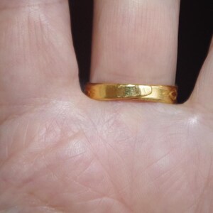 May include: A gold ring with a smooth, polished surface. The ring is worn on a finger, and the close-up shot highlights the ring's simple design and warm color. The ring appears to be made of solid gold.