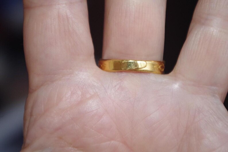 May include: A gold ring with a smooth, polished surface. The ring is worn on a finger, and the close-up shot highlights the ring's simple design and warm color. The ring appears to be made of solid gold.