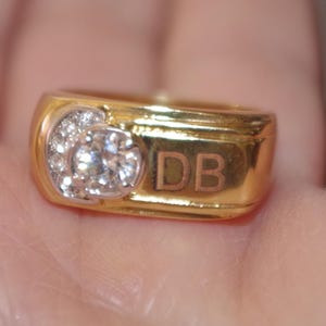 Könnte beinhalten: Ein goldener Ring mit einem großen, klaren Mittelstein und kleineren Steinen. Der Ring trägt die Initialen "DB". Ein klassisches Design, das zu verschiedenen Anlässen passt.