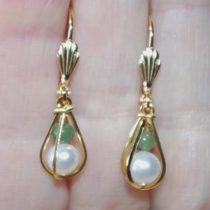 May include: A pair of gold-colored drop earrings. Each earring features a teardrop-shaped cage holding a white pearl and a green bead. The earrings have a decorative top with a fan-like design.