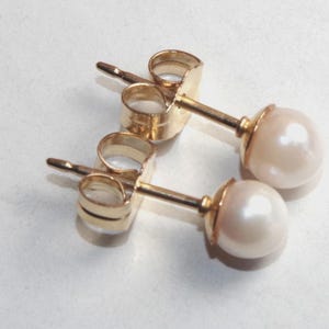 Op de afbeelding: Een paar goudkleurige oorstekers met een parel. De oorbellen hebben een pin en een vlindersluiting. De parels zijn crèmewit van kleur en rond van vorm. De oorbellen staan tegen een witte achtergrond.
