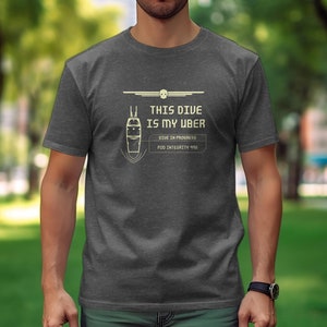 Può includere: T-shirt grigio scuro con la scritta bianca "THIS DIVE IS MY UBER". Sotto, un diagramma di una capsula subacquea e le parole "DIVE IN PROGRESS" e "POD INTEGRITY 99%."