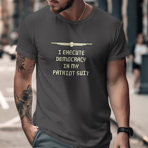 Puede incluir: Una camiseta gris oscuro con un diseño gráfico amarillo. El gráfico presenta una calavera con alas y el texto "Ejecuto la democracia en mi traje de patriota."