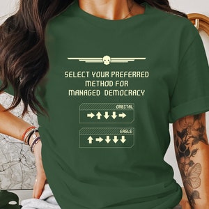 May include: A green t-shirt with a retro video game-style graphic that says "Select your preferred method for managed democracy." The graphic has two buttons, one labeled "Orbital" and the other labeled "Eagle".