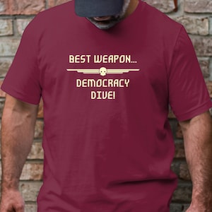 Puede incluir: Una camiseta granate con el texto "BEST WEAPON... DEMOCRACY DIVE!" en blanco. El texto está en una fuente pixelada y está rodeado por una línea blanca.