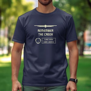 Puede incluir: Una camiseta azul marino con el texto "Remember the Creek" y un gráfico de una nave espacial con el texto "Planet Status:  A New Liberated" debajo.