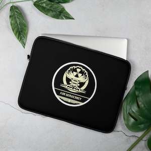 May include: Black laptop sleeve featuring a circular design with a mushroom cloud and a radiation symbol. The words "FOR DEMOCRACY" are printed below the design in a sans-serif font. The sleeve is on a light gray surface with green leaves.