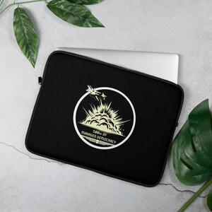 May include: Black laptop sleeve featuring a white circular graphic with two fighter jets and an explosion design. The text "500kg of Managed Democracy" is printed below the graphic. The sleeve is designed to protect a laptop.