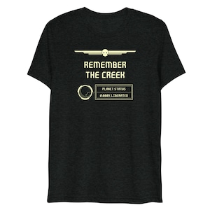 Puede incluir: Una camiseta gris oscuro con un diseño gráfico que presenta una calavera con alas, el texto "Remember the Creek" y una pantalla digital que dice "Planet Status 0.000% Liberated".