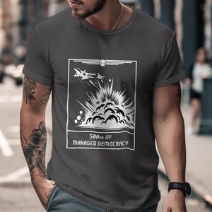 Puede incluir: Camiseta gris oscuro con un gráfico blanco de dos aviones de combate y una explosión. El gráfico incluye el texto "500hg of Managed Democracy" en un marco rectangular. La camiseta tiene mangas cortas y cuello redondo.