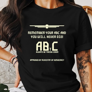 Puede incluir: Camiseta negra con un diseño gráfico blanco que dice "Remember your ABC and you will never die! AB.C Always be taking cover. Approved by Ministry of Democracy."