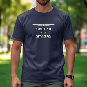 Puede incluir: Camiseta azul marino con el texto "I SPILL OIL FOR DEMOCRACY" en blanco, con un estilo retro. Encima del texto hay una línea horizontal con una calavera en el centro. La camiseta es de manga corta.