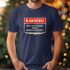 Puede incluir: Una camiseta azul marino con un cartel de advertencia que dice "¡ADVERTENCIA! PUEDE COMENZAR A HABLAR SOBRE DEMOCRACIA GESTIONADA".