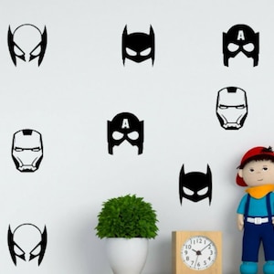 May include: Black superhero mask decals in various designs, including Wolverine, Batman, Captain America, and Iron Man. The decals are arranged on a white wall, with a clock, a plant, and a doll in the scene.