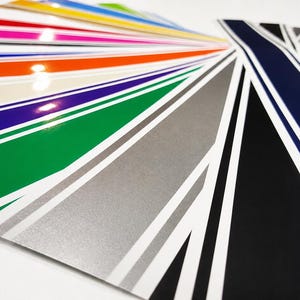 Peut inclure: Une collection d'autocollants en vinyle brillant disposés en éventail. Les bandes présentent diverses couleurs unies, notamment orange, rose, bleu, vert, argent, noir et marine, chacune accentuée par de fines rayures blanches parallèles.