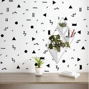 Op de afbeelding: Een witte muur met zwarte geometrische vormen en een set van drie witte geometrische wandplanters. De planters hangen aan de muur en zijn gevuld met groene planten.