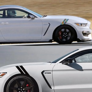 Può includere: Due Ford Mustang bianche con strisce da corsa nere sui parafanghi anteriori. L'auto in alto ha una striscia arcobaleno sul parafango.