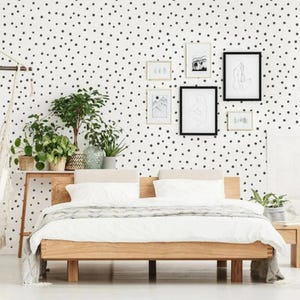 May include: A modern bedroom with a wooden bed frame, white bedding, and a gray blanket. The wall is decorated with black polka dots and framed artwork. Potted plants and a wooden side table add to the room's decor.