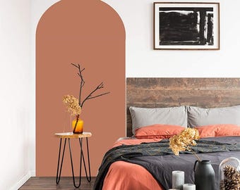 Puede incluir: Escena de dormitorio con estética moderna. Una cama con ropa de cama coral y gris en primer plano. Una pequeña mesa redonda con patas de horquilla negras sostiene un jarrón con flores secas. Un arco pintado en color terracota está en la pared.