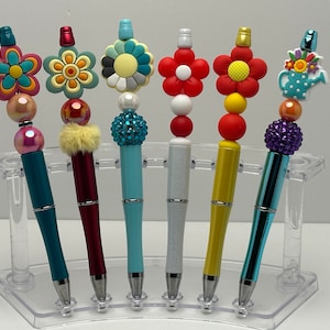 May include: Six colorful pens with different flower and watering can designs on top. The pens are displayed on a clear plastic stand.