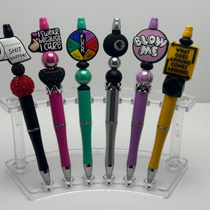 May include: Six colorful pens with different decorative tops. The tops include a toilet paper roll, a hand gesture, a spinning wheel, a billiard ball, a pink circle, and a yellow sign. The pens are displayed on a clear plastic stand.