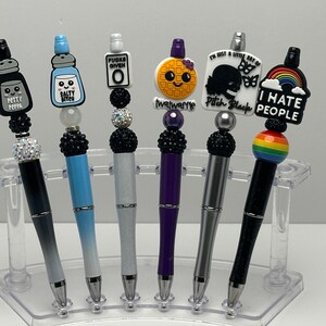 May include: Six decorative pens with different colored bodies and tops. The tops feature various designs and sayings, including "Petty Peppa", "Salty Bitch", "Fucks Given 0", "I'm Just A Little Bit Of Twatwarme", "Pitch Black", and "I Hate People".